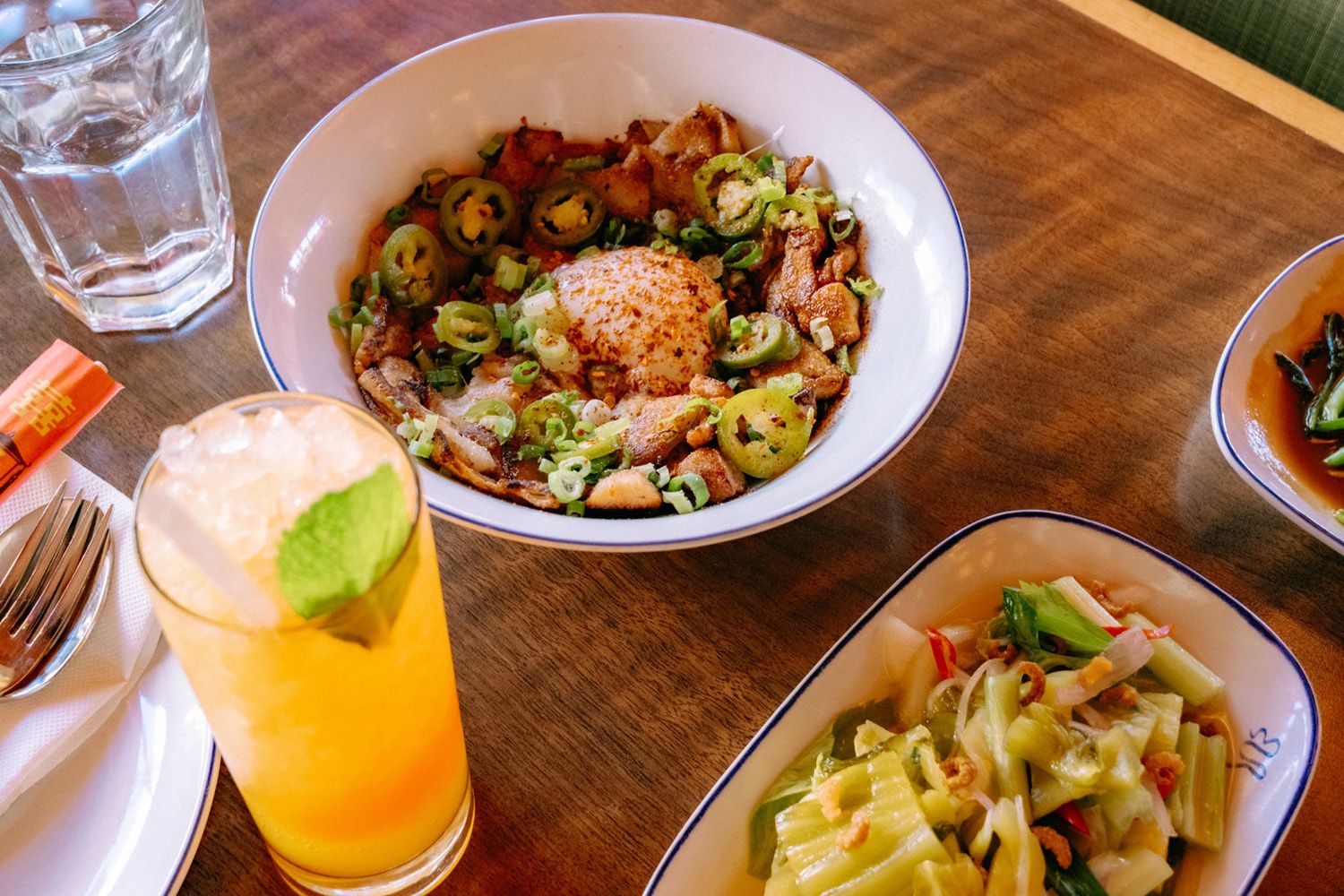 A bowl of spicy noodles with meat, a glass of orange juice, a water glass, cutlery, and a plate of vegetables on a wooden table