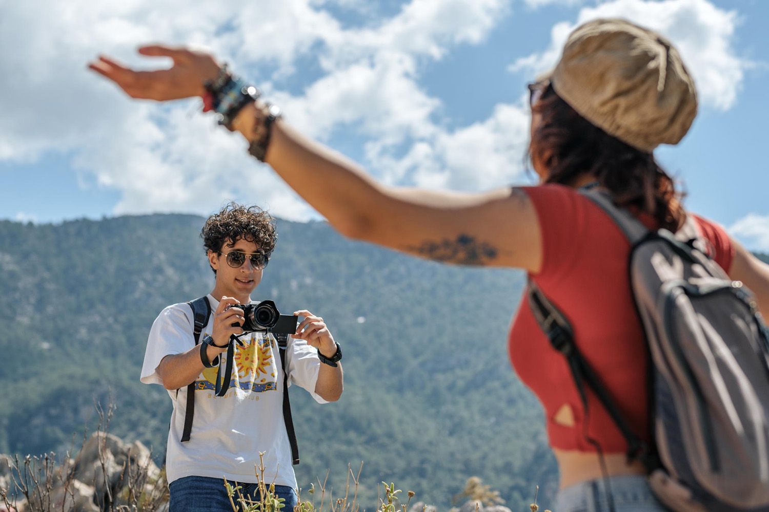 A person holds a camera and smiles at another with arms raised, set against a scenic mountain backdrop under a partly cloudy sky