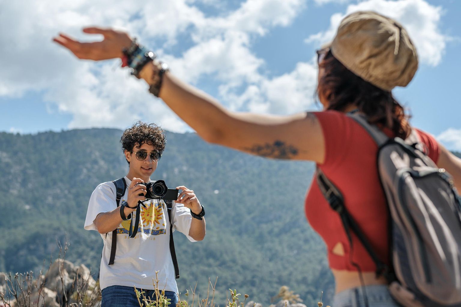 A person holds a camera and smiles at another with arms raised, set against a scenic mountain backdrop under a partly cloudy sky