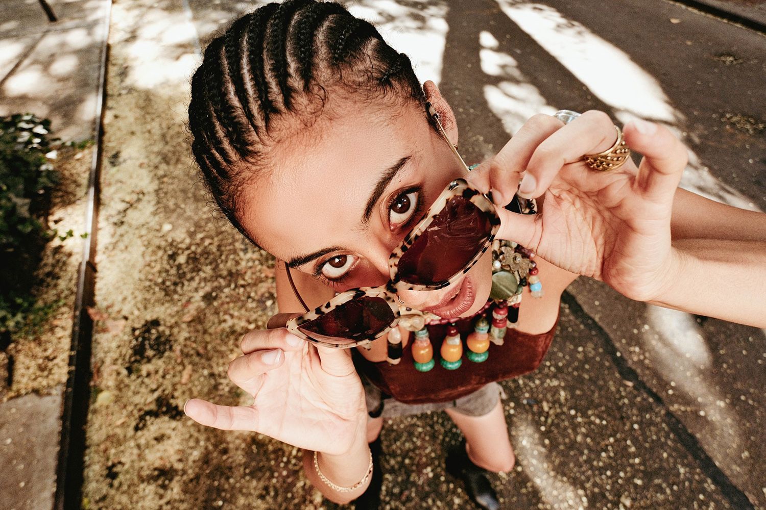 Person holds sunglasses, looking playfully at the camera on a sunny street
