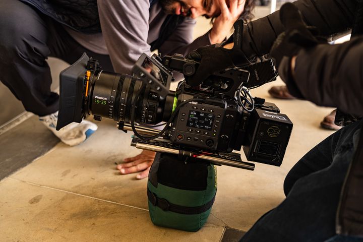 A professional film camera is set up on a cushioned support on the floor, with two people adjusting equipment nearby