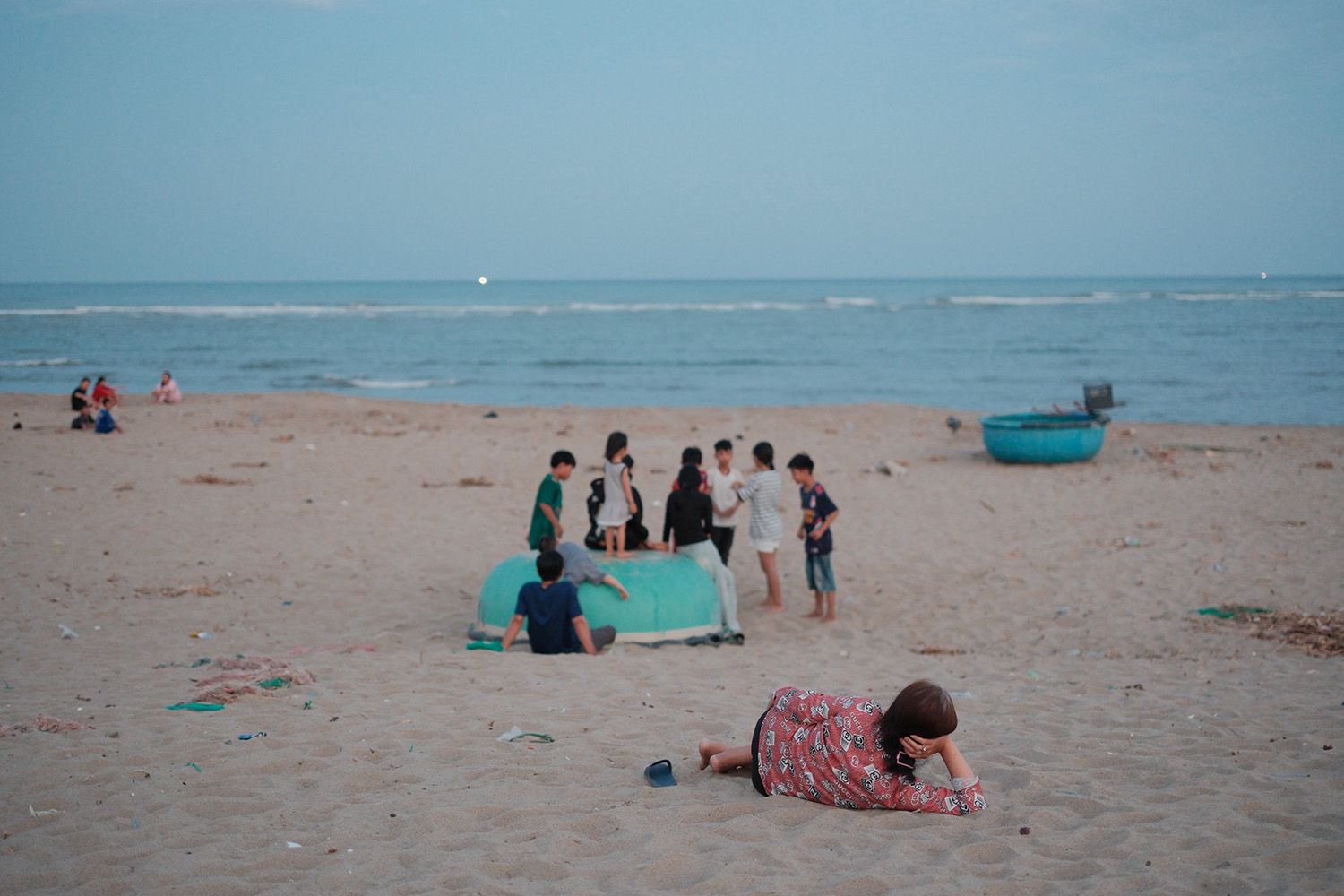 Person lying on a sandy beach, watching a group of people gathered near a turquoise boat with the ocean in the background