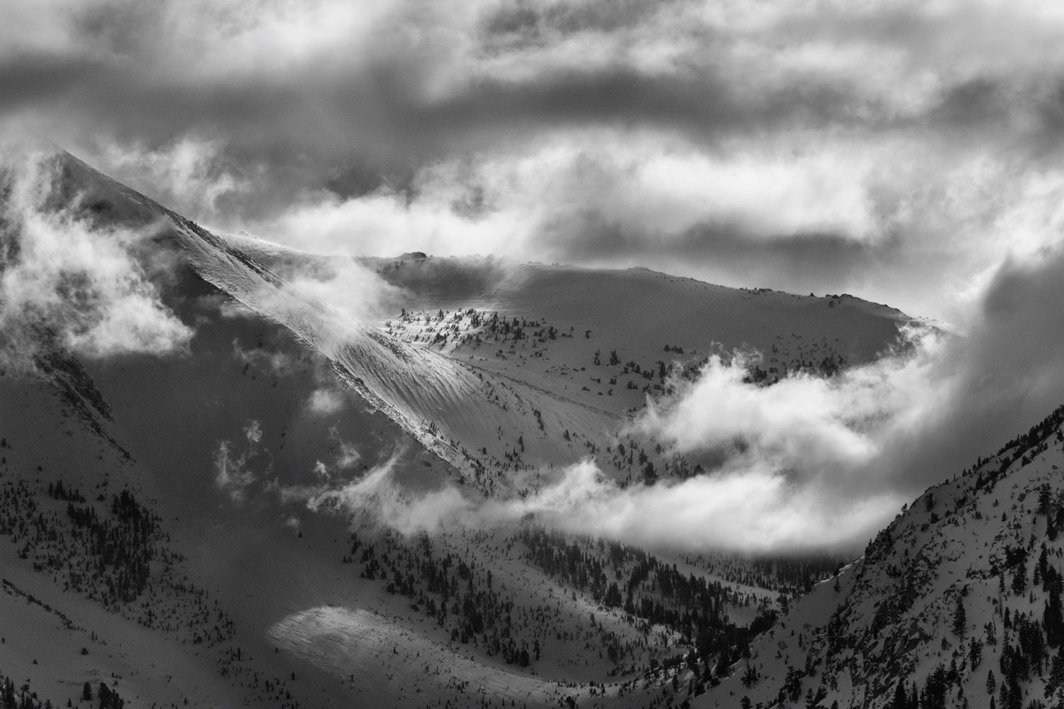 Snow-covered mountains partially shrouded in mist, with clouds drifting across the rugged peaks under a moody sky