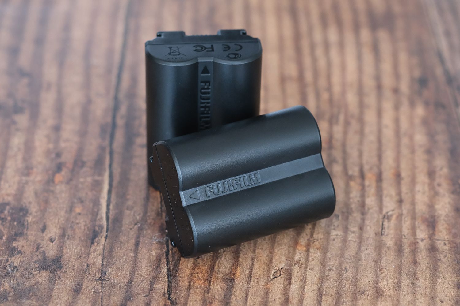 Two black Fujifilm camera batteries are placed upright on a wooden surface, one leaning against the other