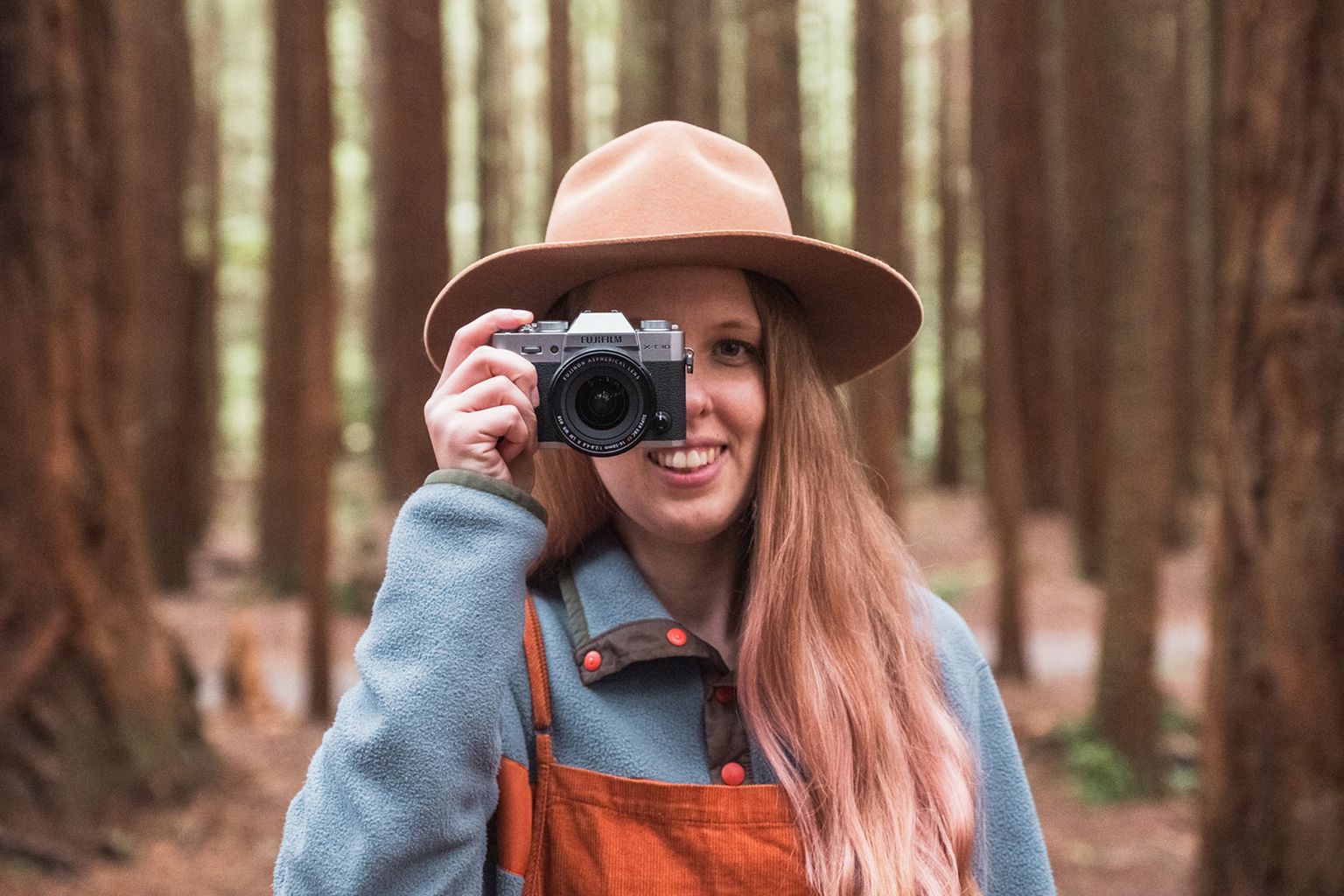 Person with long hair and hat smiles, holding a camera in a forest with tall trees