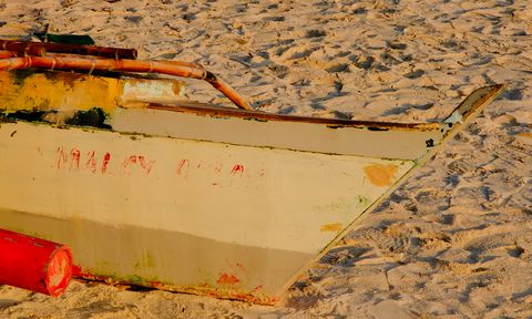 A close-up of a weathered wooden boat on sandy shore, with visible peeling paint and worn textures in soft, warm light