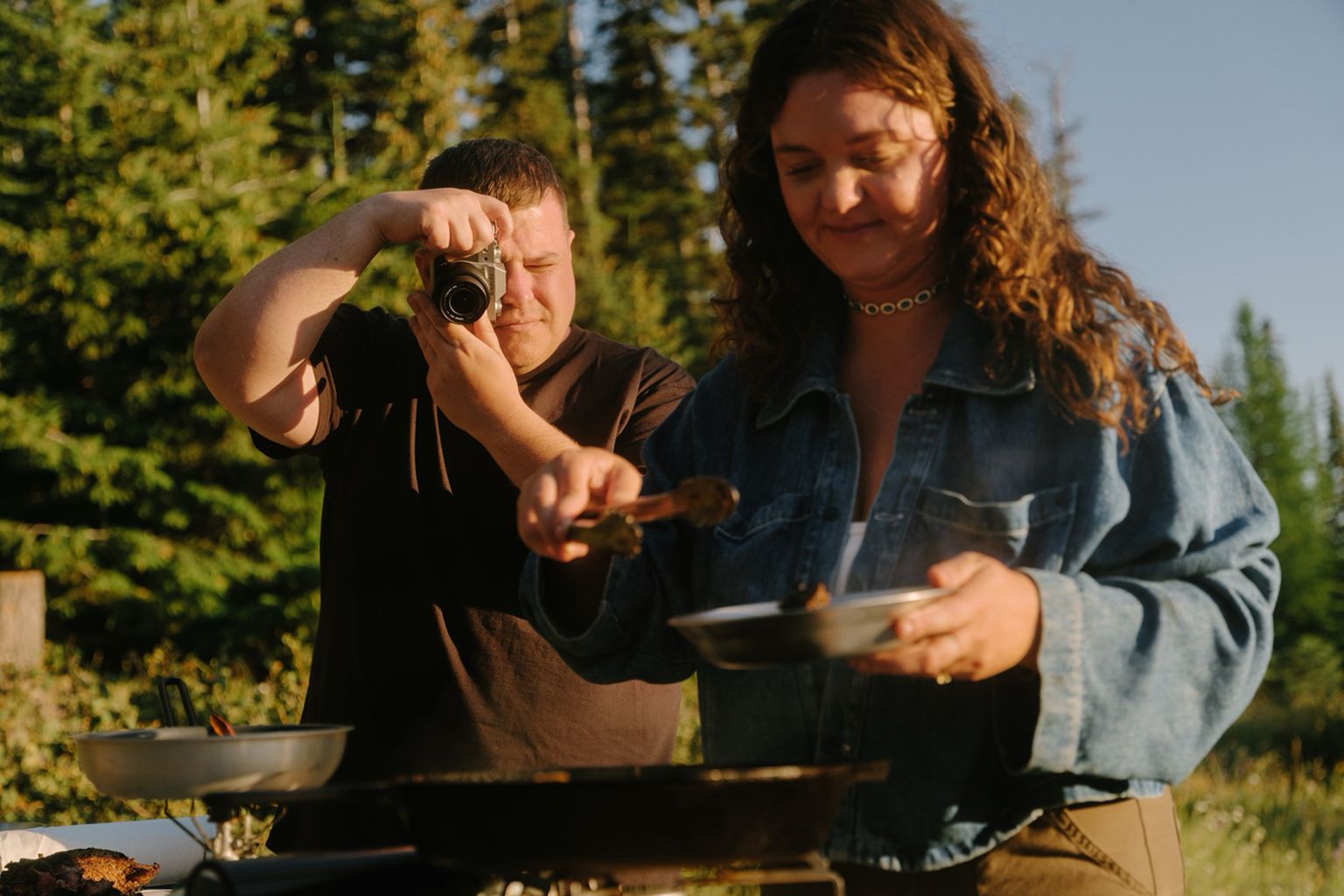 Two people outdoors; one is photographing, the other cooking with a skillet, surrounded by trees under a clear sky