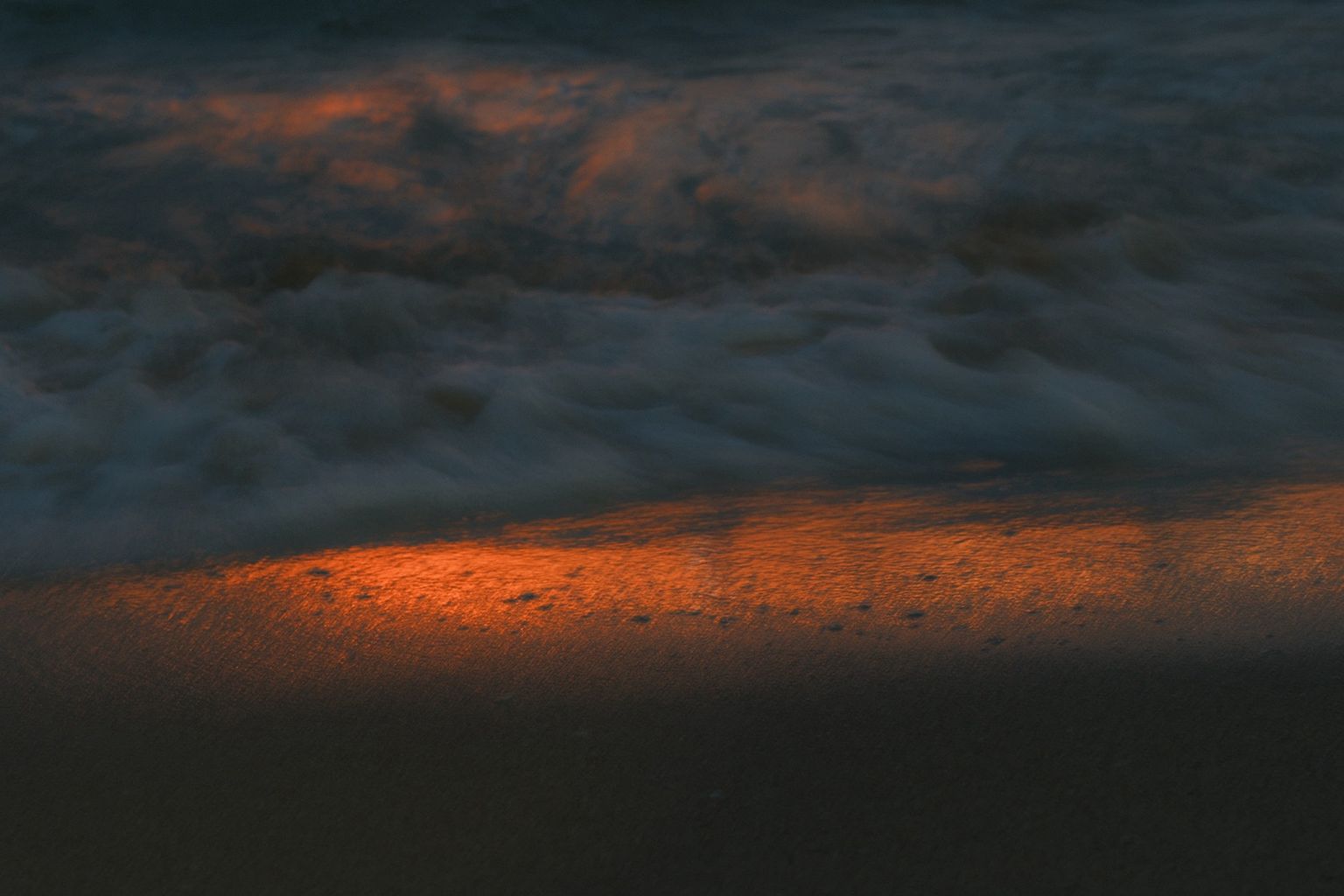 Gentle waves wash over sand at dusk, illuminated by warm, orange light reflecting on the shore
