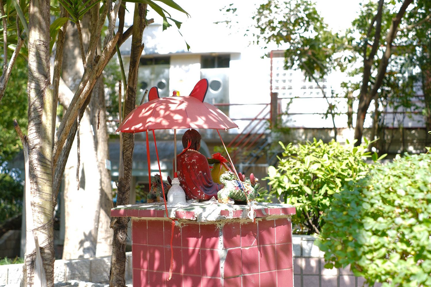 A small stall with a red tiled structure, featuring a statue under a red umbrella, surrounded by green foliage and trees