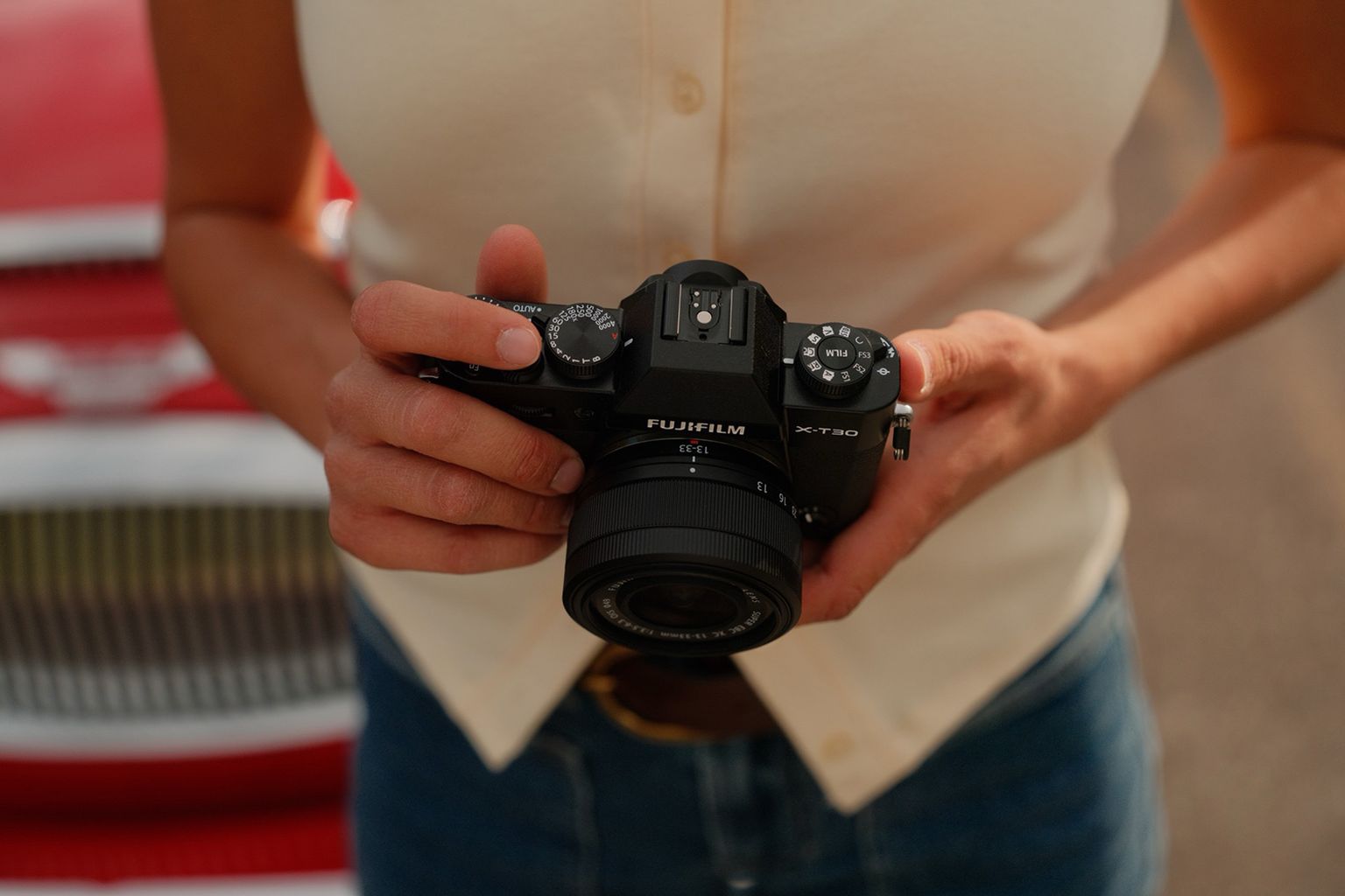 Person holding FUJIFILM X-T30 III, standing in front of a red and white vintage car, wearing a beige top and blue jeans