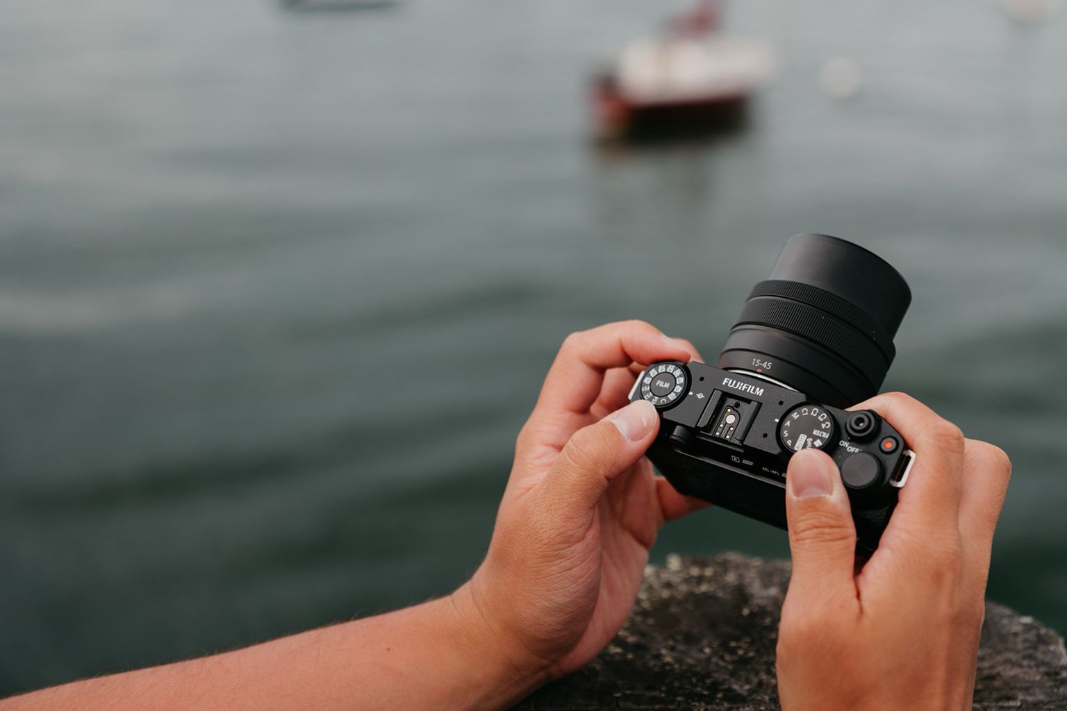 Hands holding a camera, focused on adjusting settings, with a blurred body of water and a distant boat in the background
