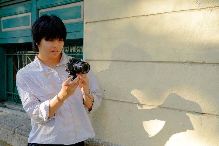 Person with short dark hair holding a camera, standing by a sunlit wall