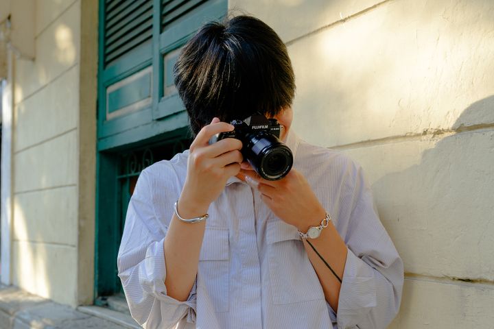Person with black hair using a camera, leaning against a wall in sunlight, wearing a light-colored shirt