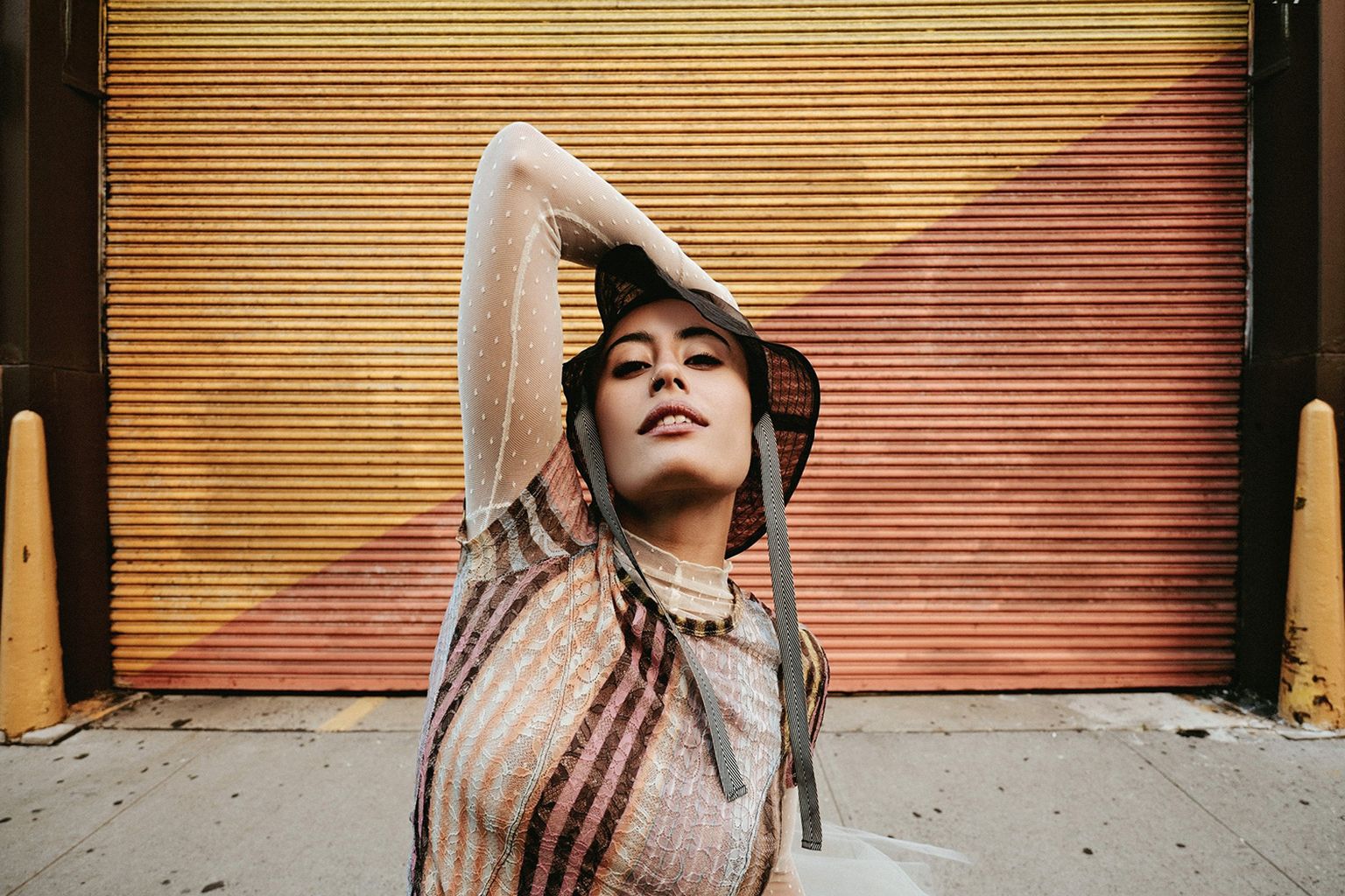 Person posing with arm above head, wearing a patterned top and hat, stands in front of a colorful corrugated metal door on a sidewalk
