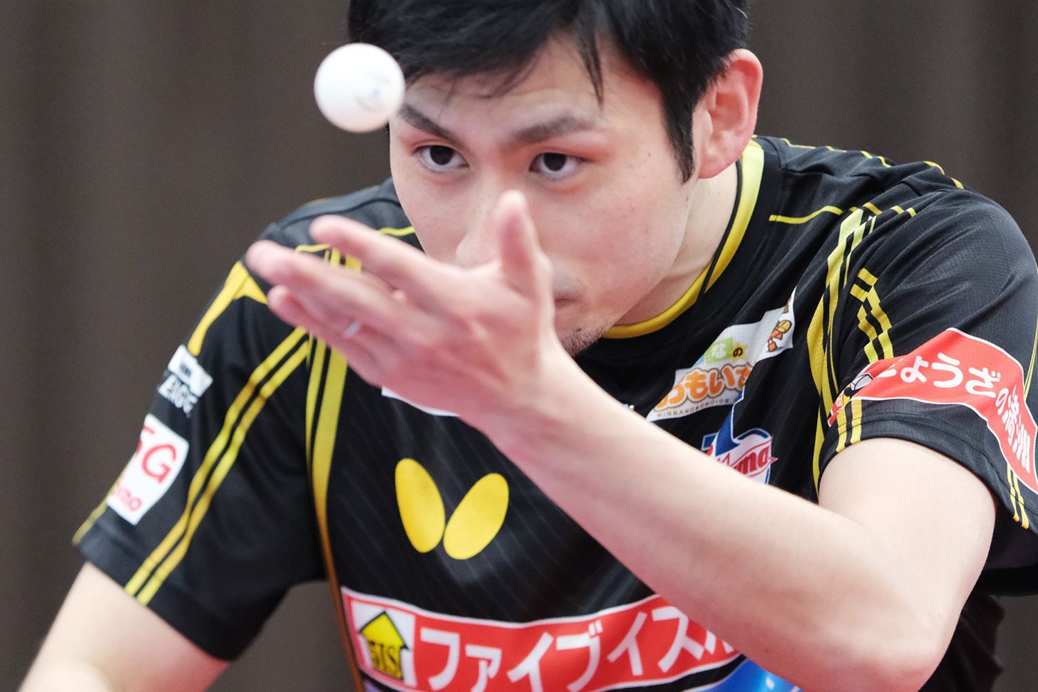 A table tennis player in motion, concentrating intensely as he prepares to serve, with a ball hovering near his face