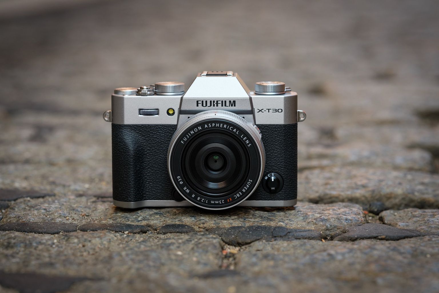 FUJIFILM X-T30 III placed on a cobblestone ground, showing its front view with lens and dials visible