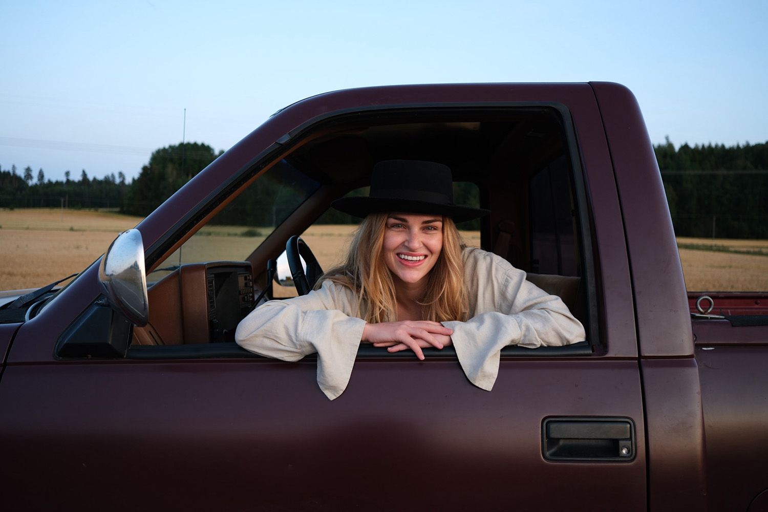 A smiling person in a hat leans out of a brown truck's window, set against a rural background with fields and trees