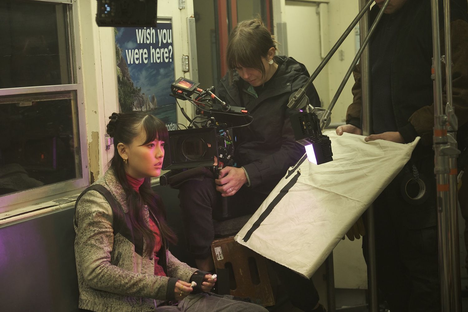Actress seated on a set, surrounded by crew with camera and lighting equipment, in a train-like setting