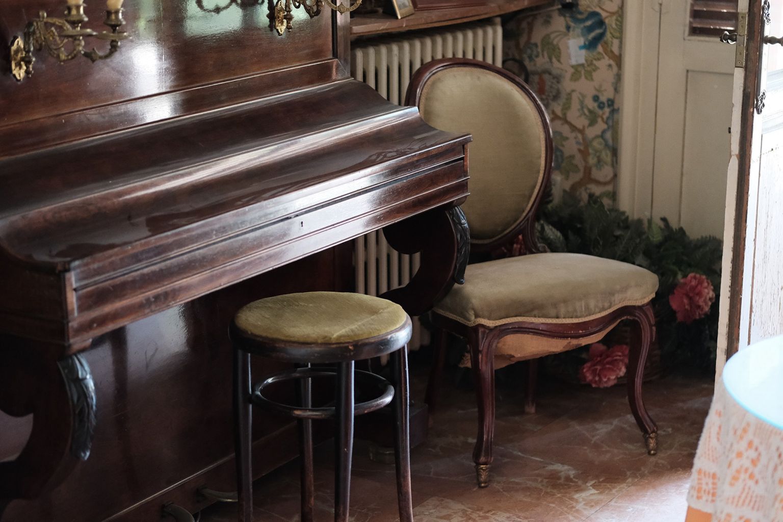 Antique wooden piano with a cushioned stool, alongside an ornate upholstered chair in a cozy, vintage room