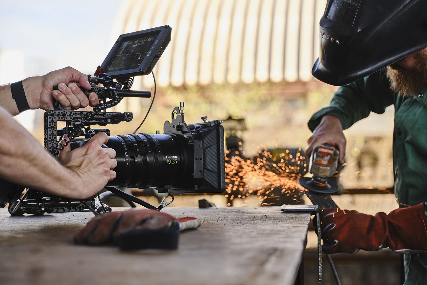 Person grinding metal with sparks flying, while a camera operator films closely on a wooden surface in an industrial setting