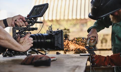 Person grinding metal with sparks flying, while a camera operator films closely on a wooden surface in an industrial setting