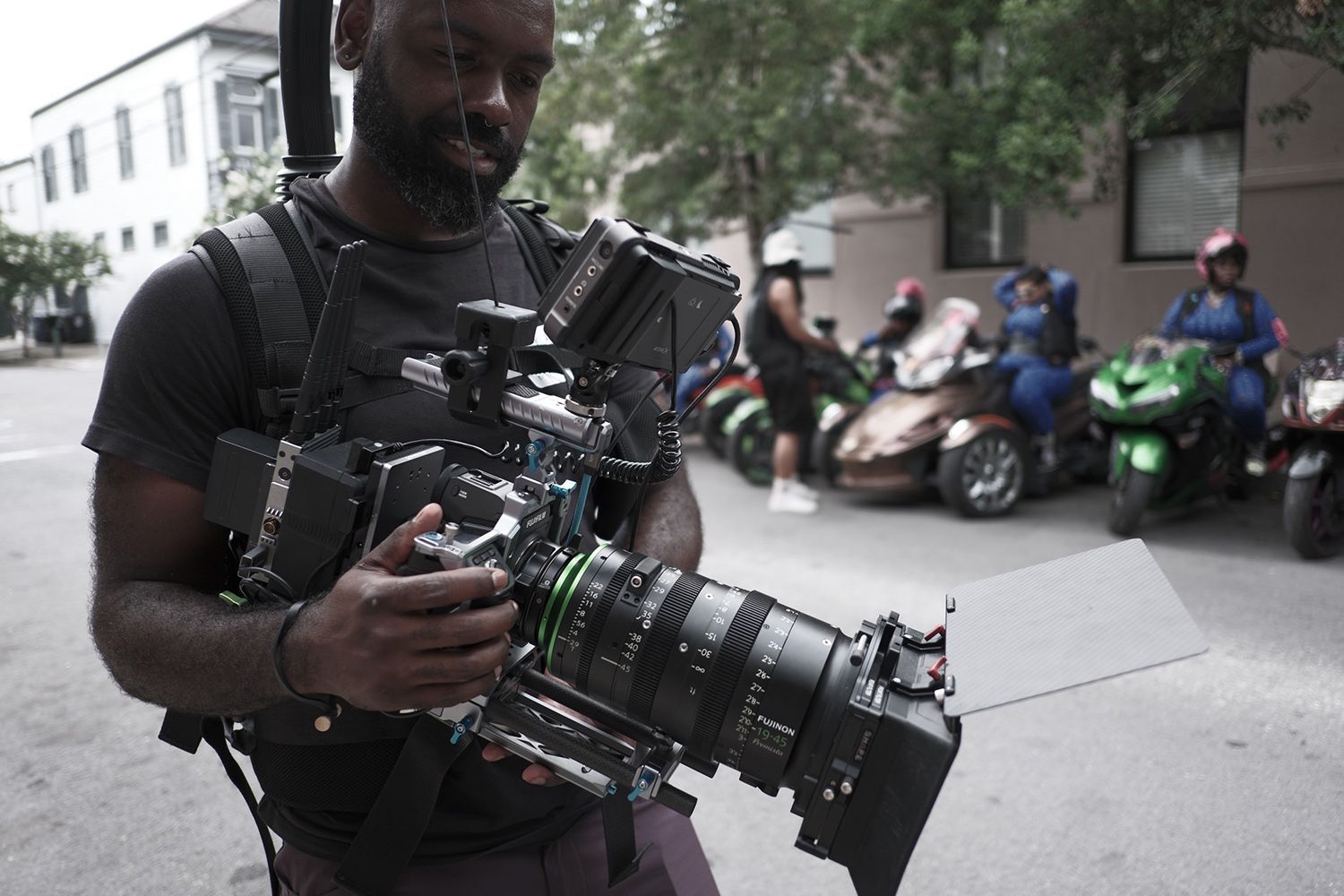 A person wearing a harness operates a professional camera on a street, with several motorcycles and riders parked in the background