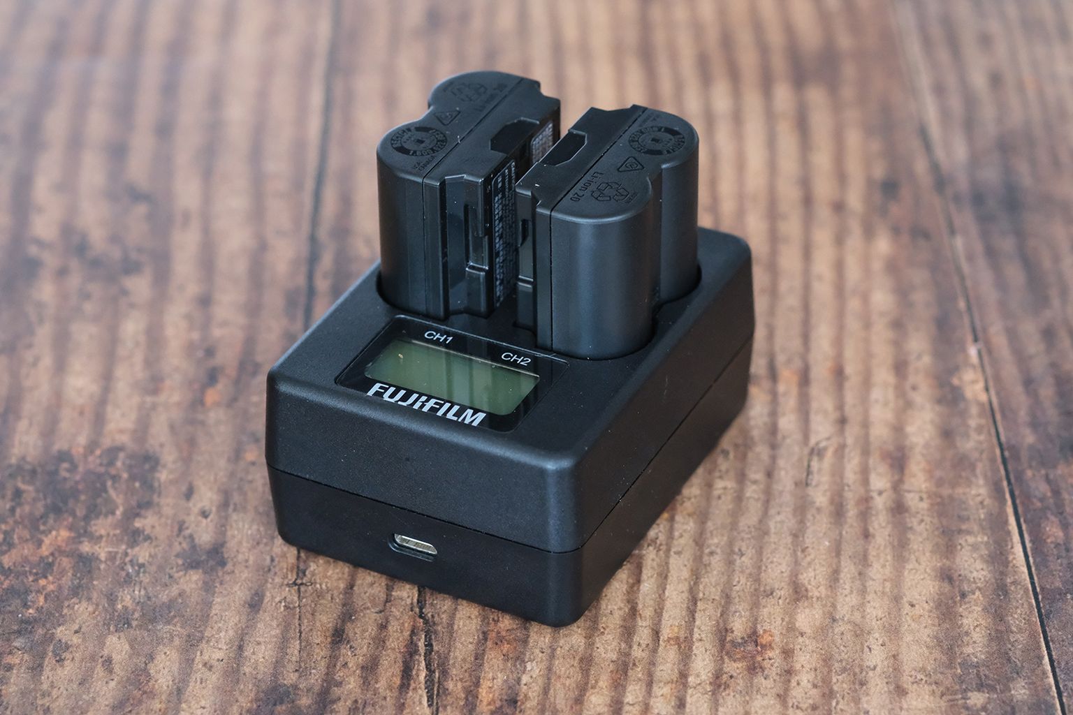 A Fujifilm battery charger with two batteries inserted, placed on a wooden surface