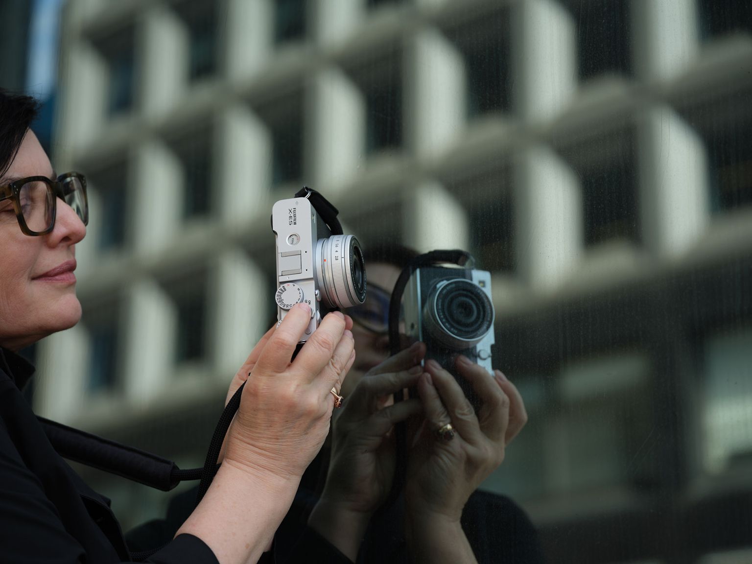 A person holds a camera close to a reflective glass surface, capturing an image. The background shows a building with a geometric surface A person holds a camera close to a reflective glass surface, capturing an image. The background shows a building with a geometric surface