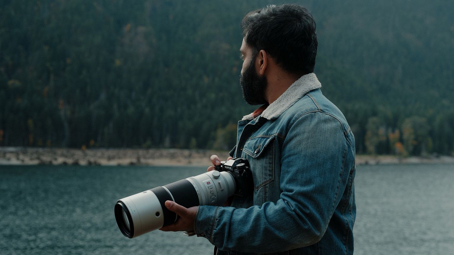 A man in a denim jacket holds a camera with a long lens, standing by a lake with a backdrop of dense forest