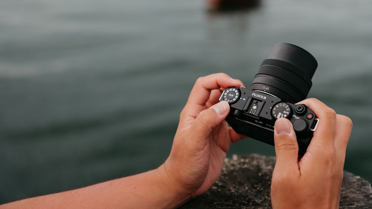 A person holds a digital camera with a large lens, adjusting the settings, with a blurred body of water is visible in the background