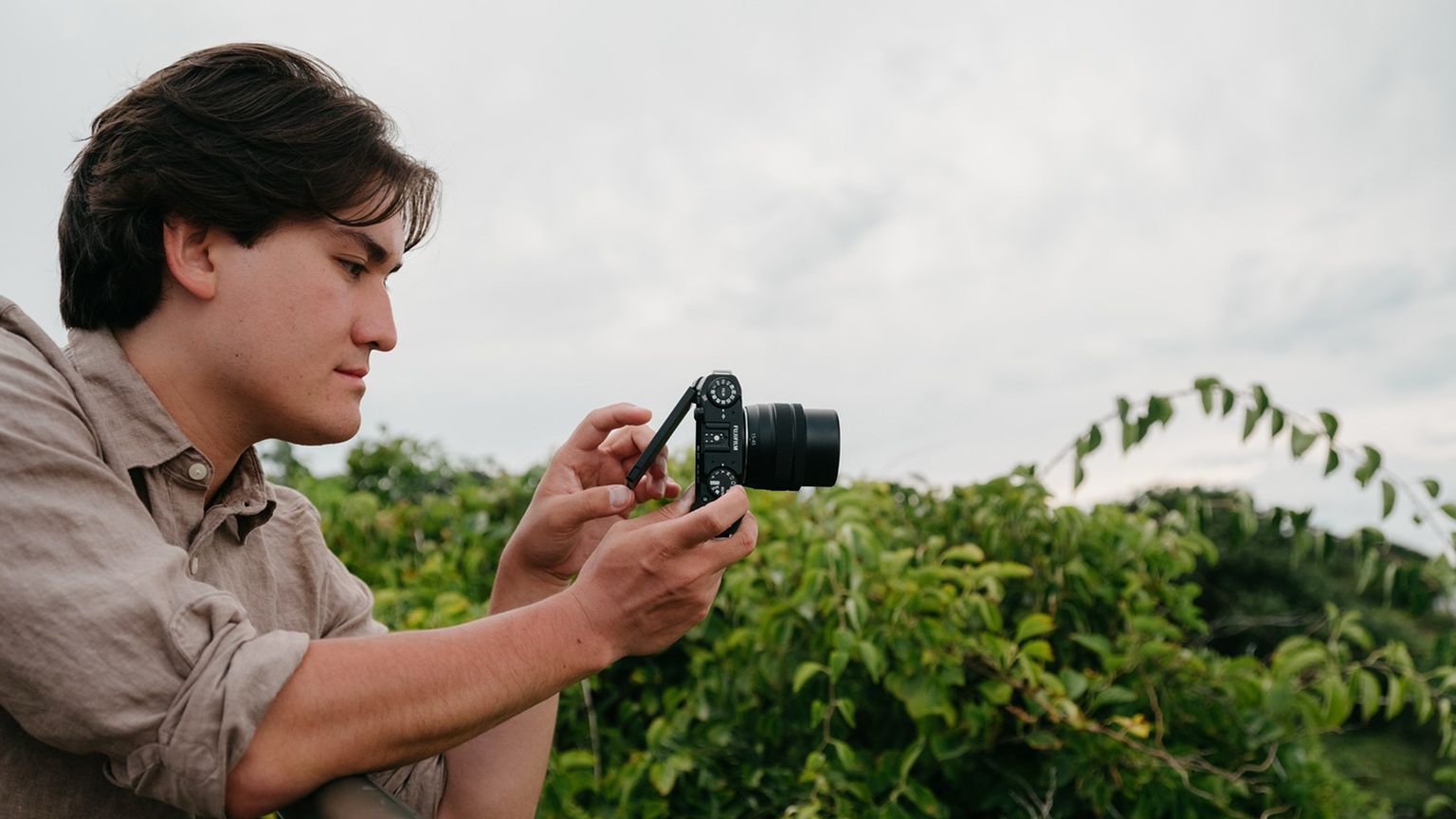 Person holding a camera, adjusting settings, with a lush green background and cloudy sky