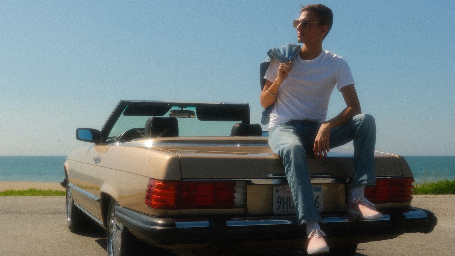 A person sits on a convertible car's trunk by the seaside, wearing a white shirt and jeans, with the sun shining brightly