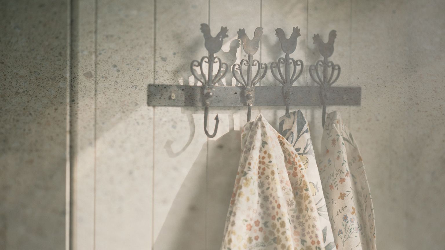 Metal hooks with rooster designs holding floral-patterned cloths on a wooden wall with a rustic ambience