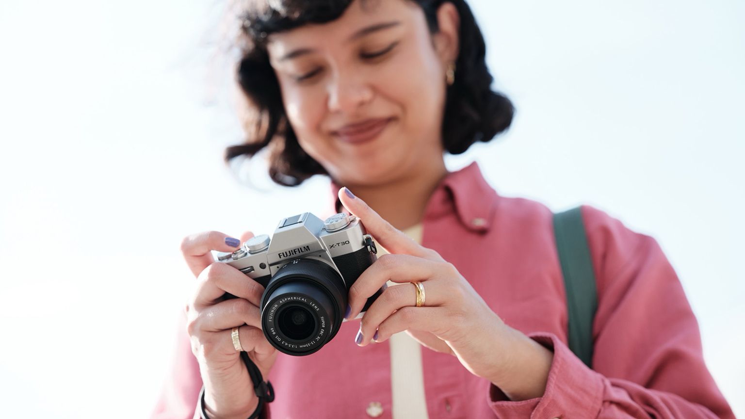 A person in a pink shirt is smiling and holding a camera, looking down at it against a bright sky background