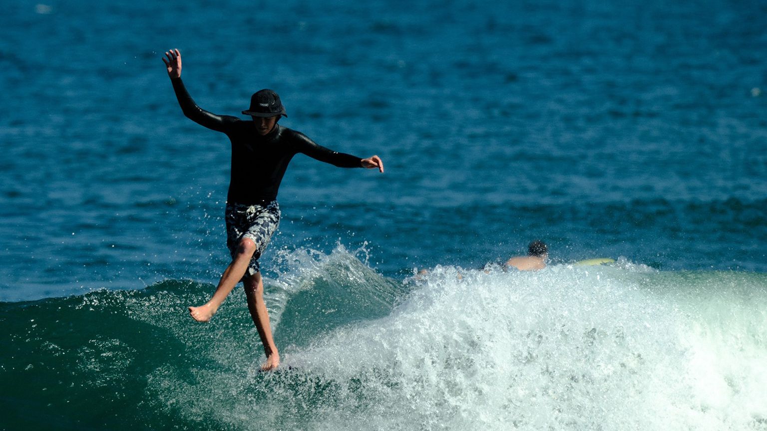 Surfer balancing on a wave, wearing a black shirt and patterned shorts, with the ocean in the background