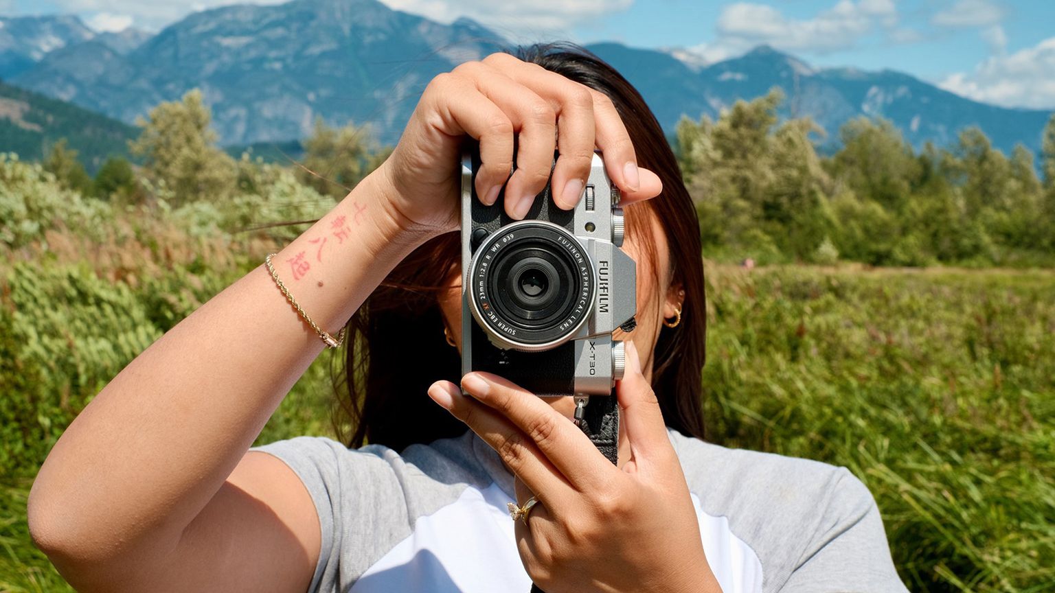 A person holding a camera to their face, standing in a grassy field with mountains and a blue sky in the background