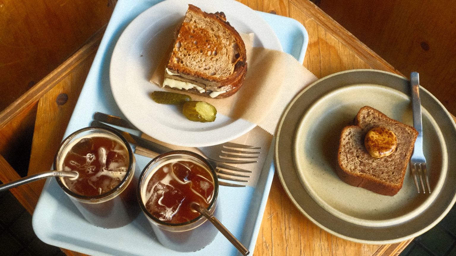 Grilled sandwich with pickles and two iced coffees on a tray, alongside a plate with a slice of bread topped with a dollop, on a wooden table