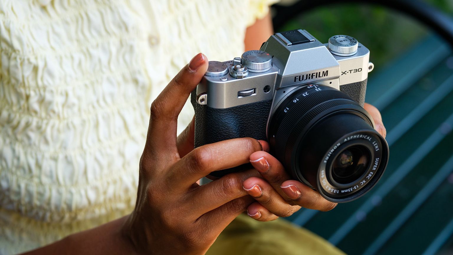 Hands holding FUJIFILM X-T30 III with a lens attached, against a background of a bench and greenery