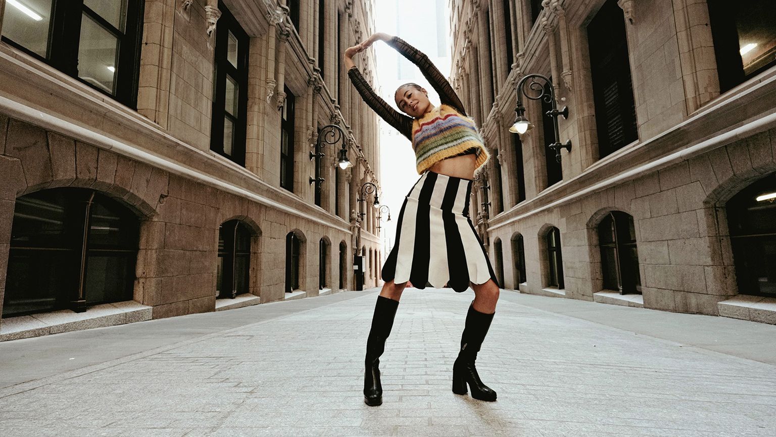 Person posing confidently in a stylish outfit in an empty urban street between tall buildings, arms raised above their head