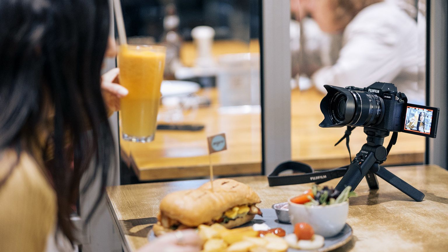 Person holding a glass of orange juice in front of a camera on a tripod, with a sandwich, fries, and salad on the table