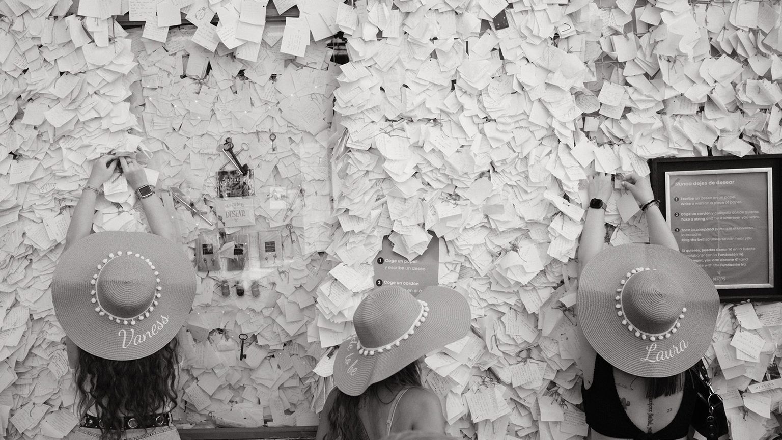 Three people in wide-brimmed hats pin notes on a wall covered with papers, in a black and white image