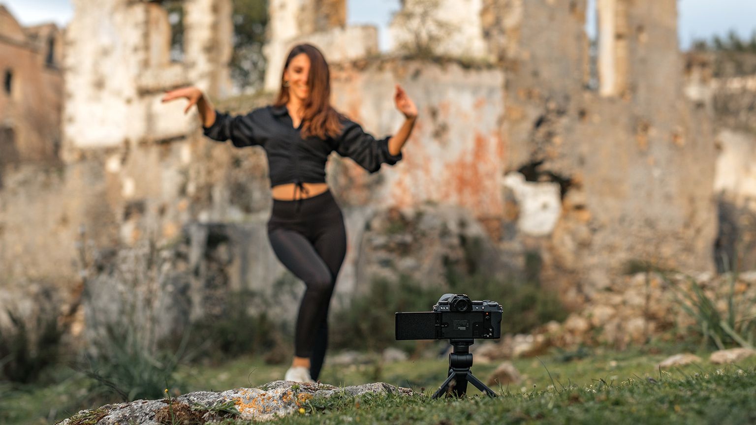 A woman dances in front of a camera on a tripod, set against the backdrop of ancient stone ruins in an outdoor setting