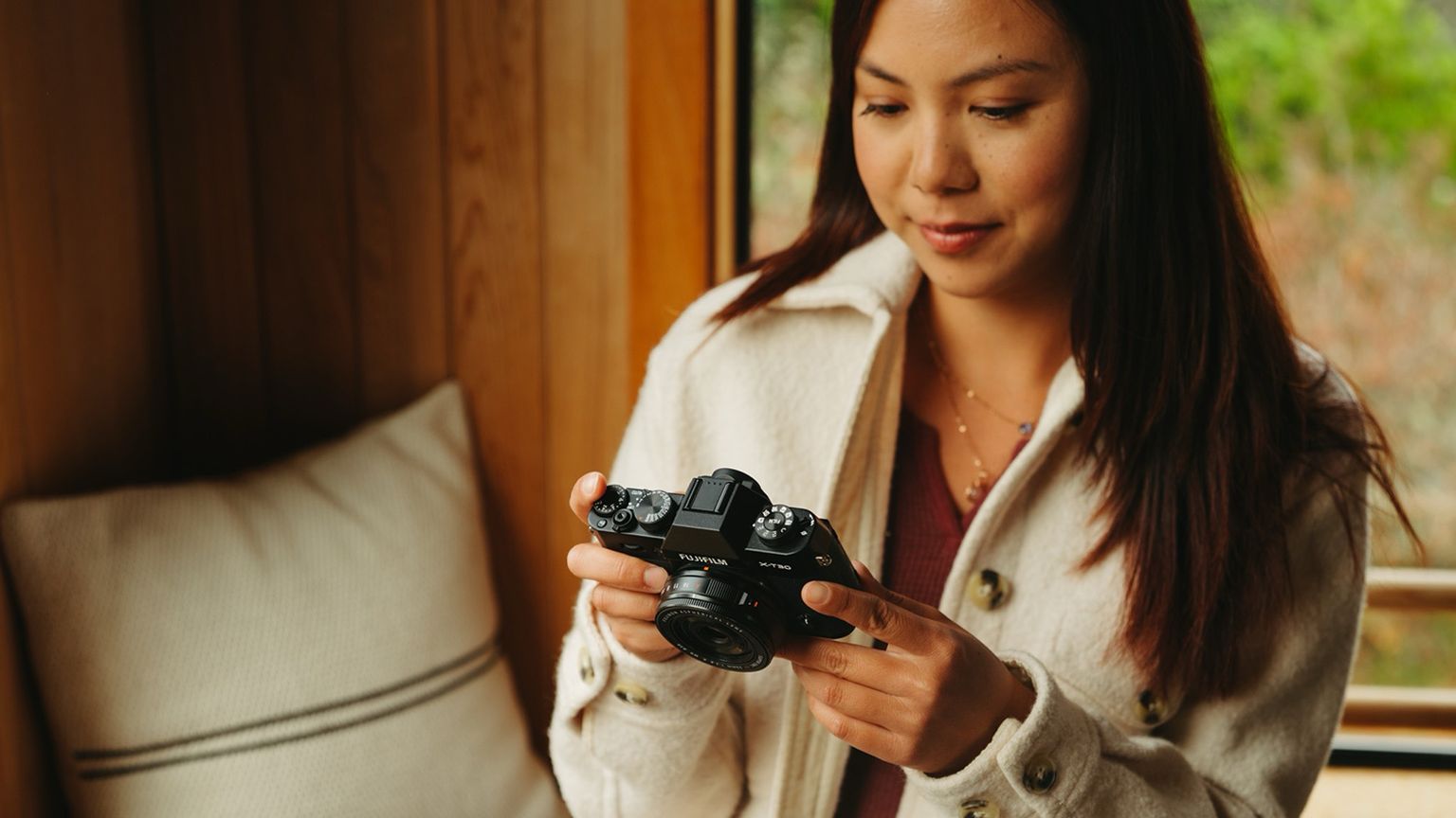 A woman in a light jacket holds a camera, standing by a window with a pillow on the bench