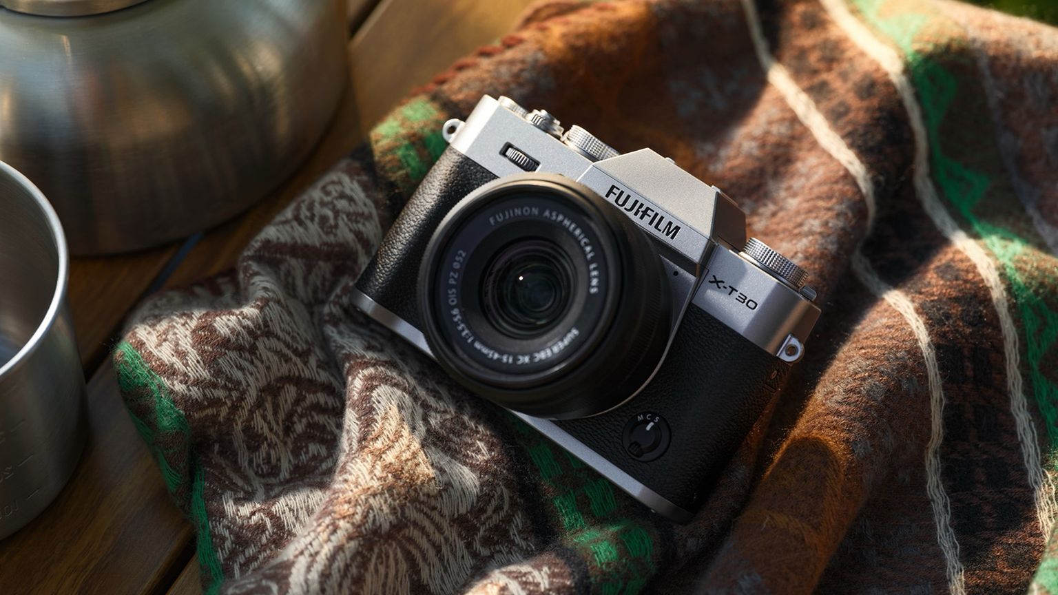 FUJIFILM X-T30 III rests on a patterned fabric surface, surrounded by a metal object and warm, natural lighting