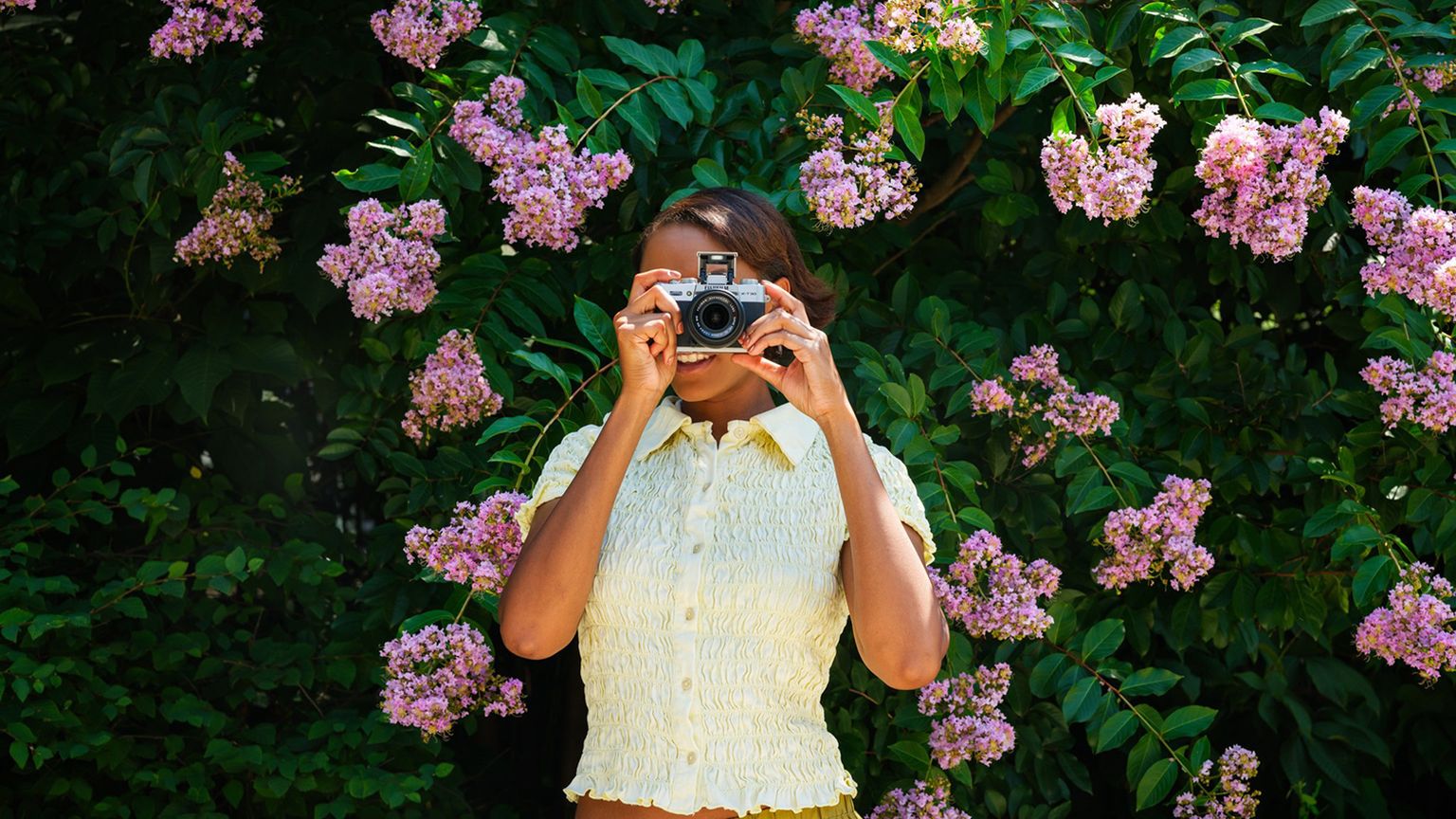 A person in a yellow top holds a camera, standing in front of lush green foliage with vibrant pink flowers