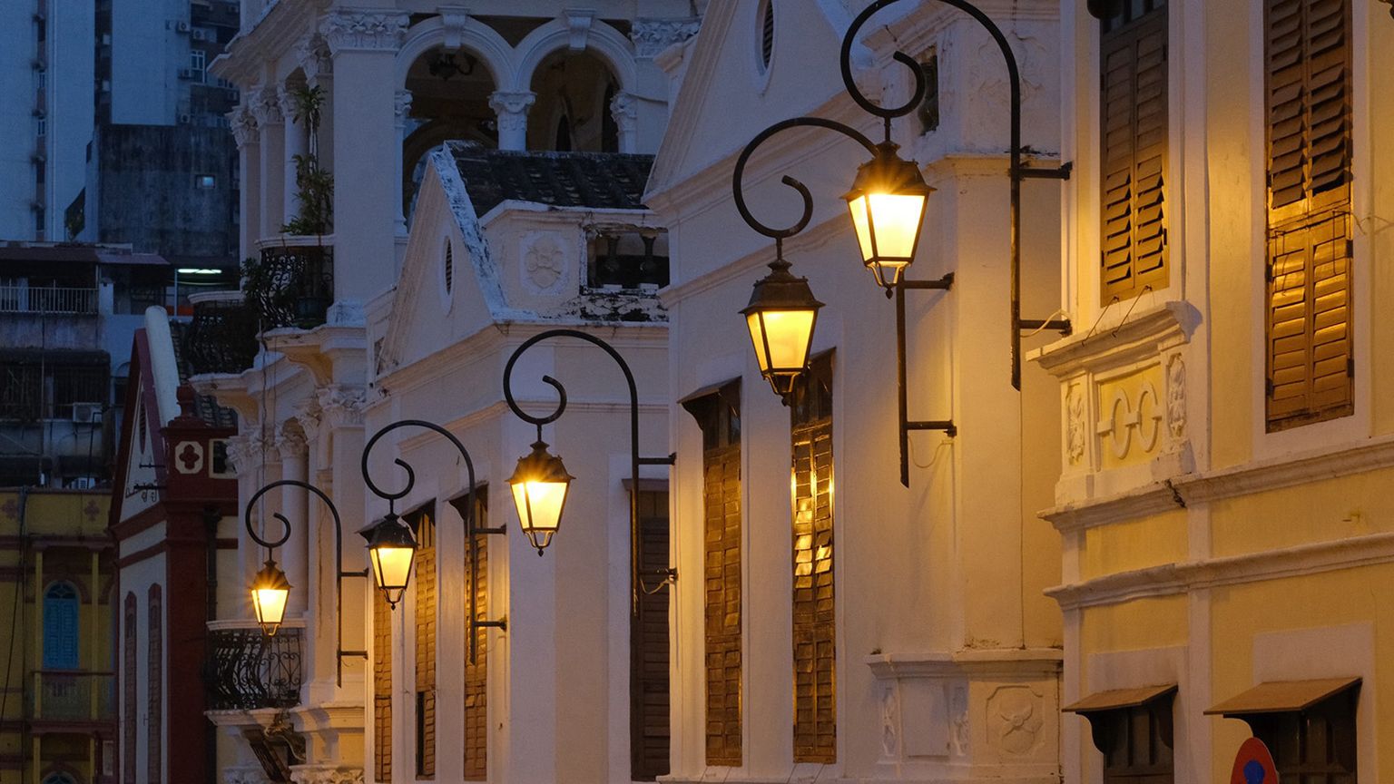 Illuminated street lamps line a row of ornate, historic buildings with shutters, creating a warm, nostalgic evening ambiance