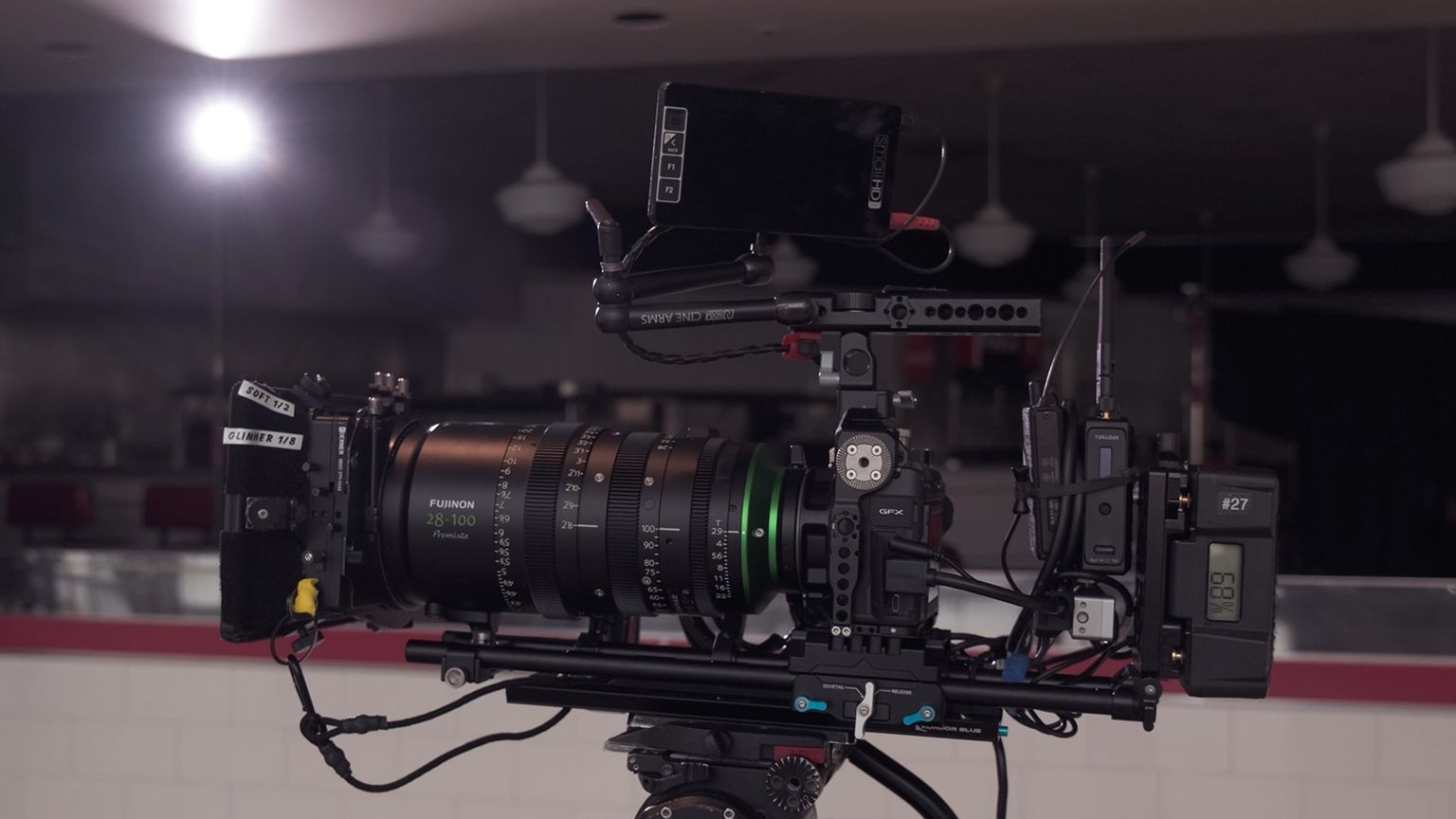 Professional video camera setup with large lens, mounted on a tripod, in a dimly lit room with ceiling lights