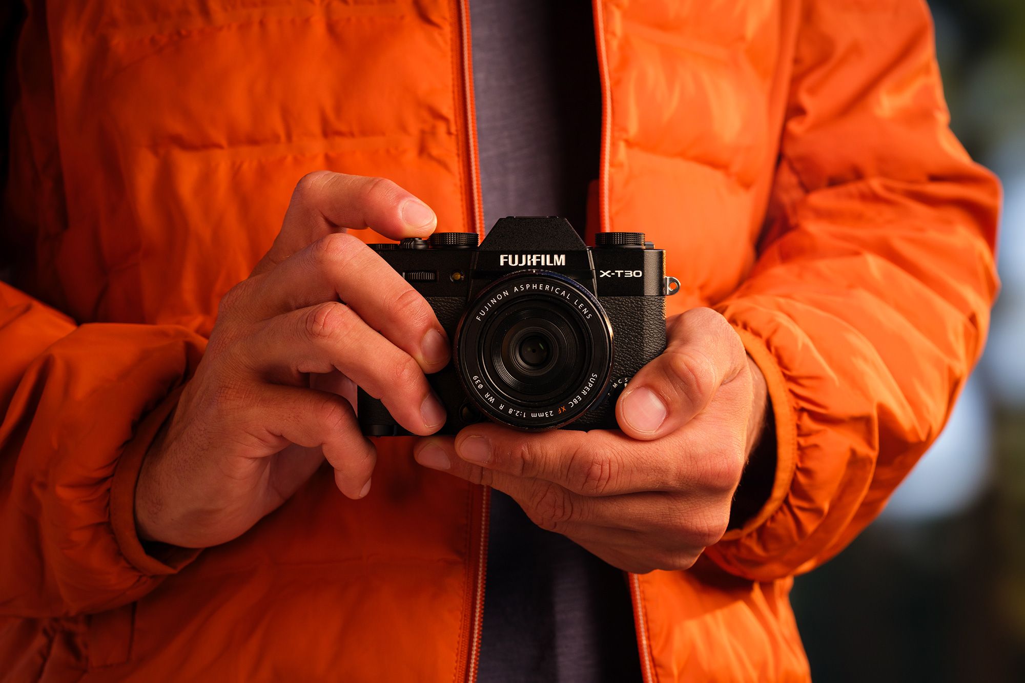 A person in an orange jacket holding FUJIFILM X-T30 III with both hands, set against a blurred background.