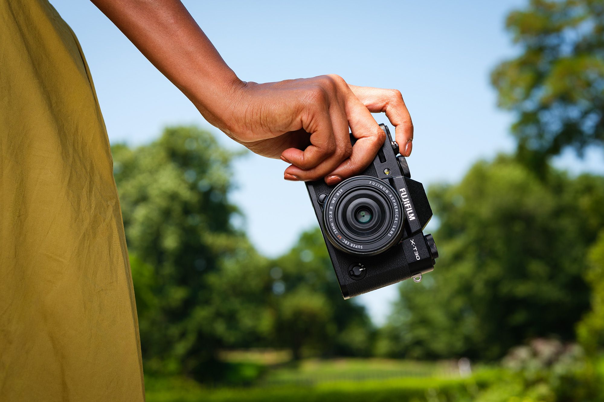 A hand holding FUJIFILM X-T30 III against a backdrop of blurred greenery and blue sky.