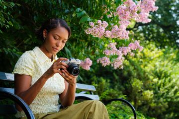 A woman in a yellow blouse sits on a bench, examining FUJIFILM X-T30 III. Pink flowers and lush greenery surround her in a sunlit garden.